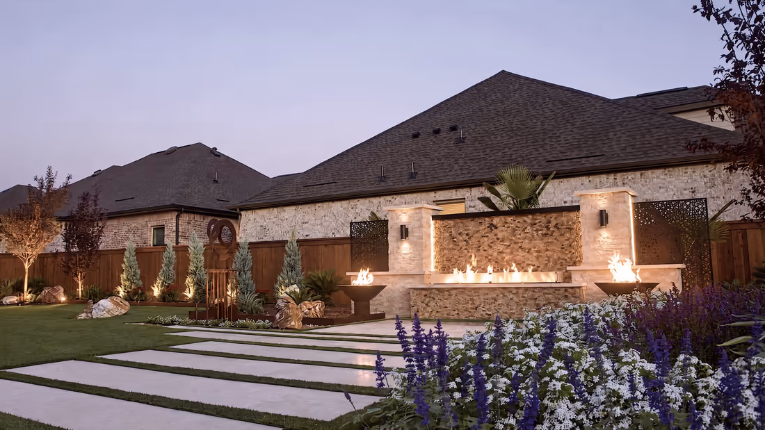 The finished yard at dusk: lit fire bowls along a stone water-and-fire wall, paver walkway and full planting beds in flower.